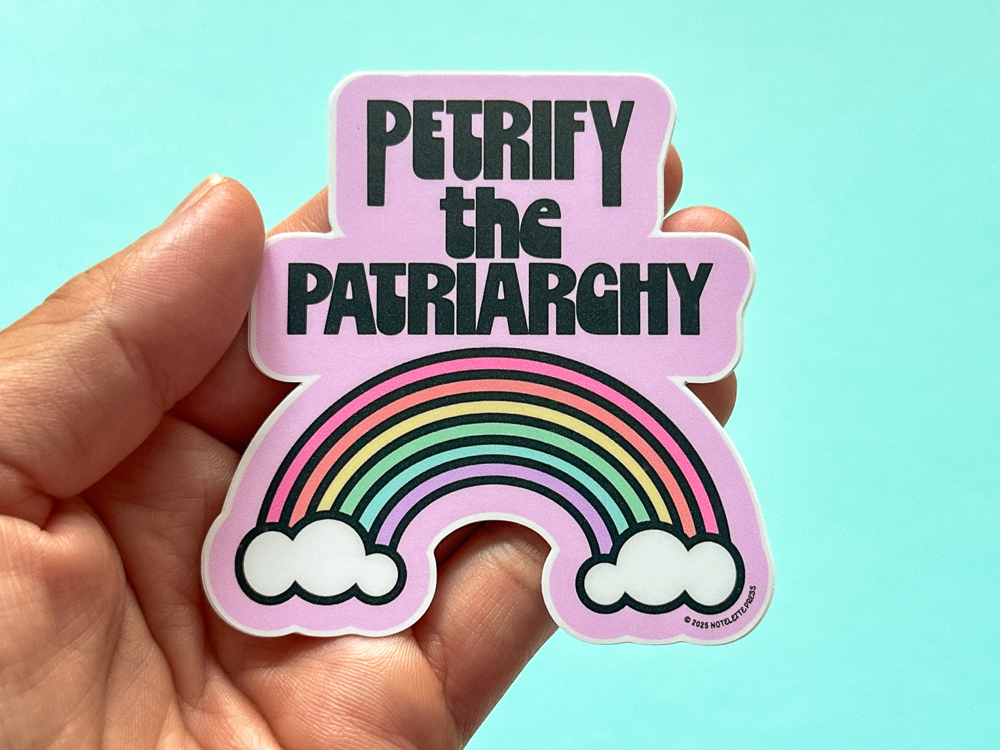 Sticker with rainbow and text 'Petrify the Patriarchy' held by a hand against a light blue background
